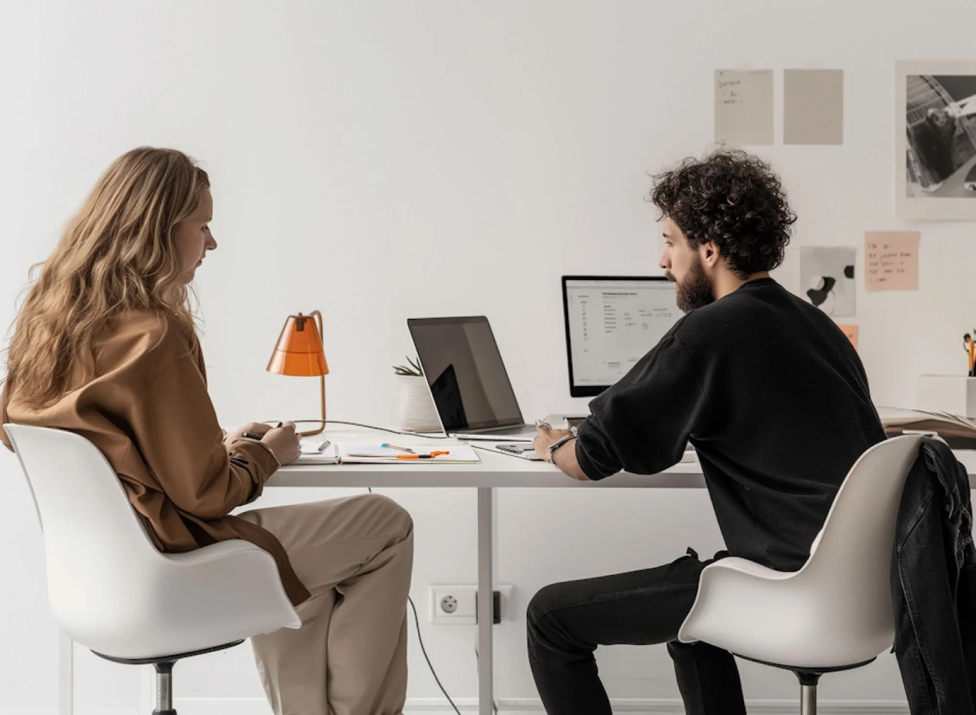 Two people sitting across from each other at a white table with laptops and papers, engaged in discussion in a modern office.